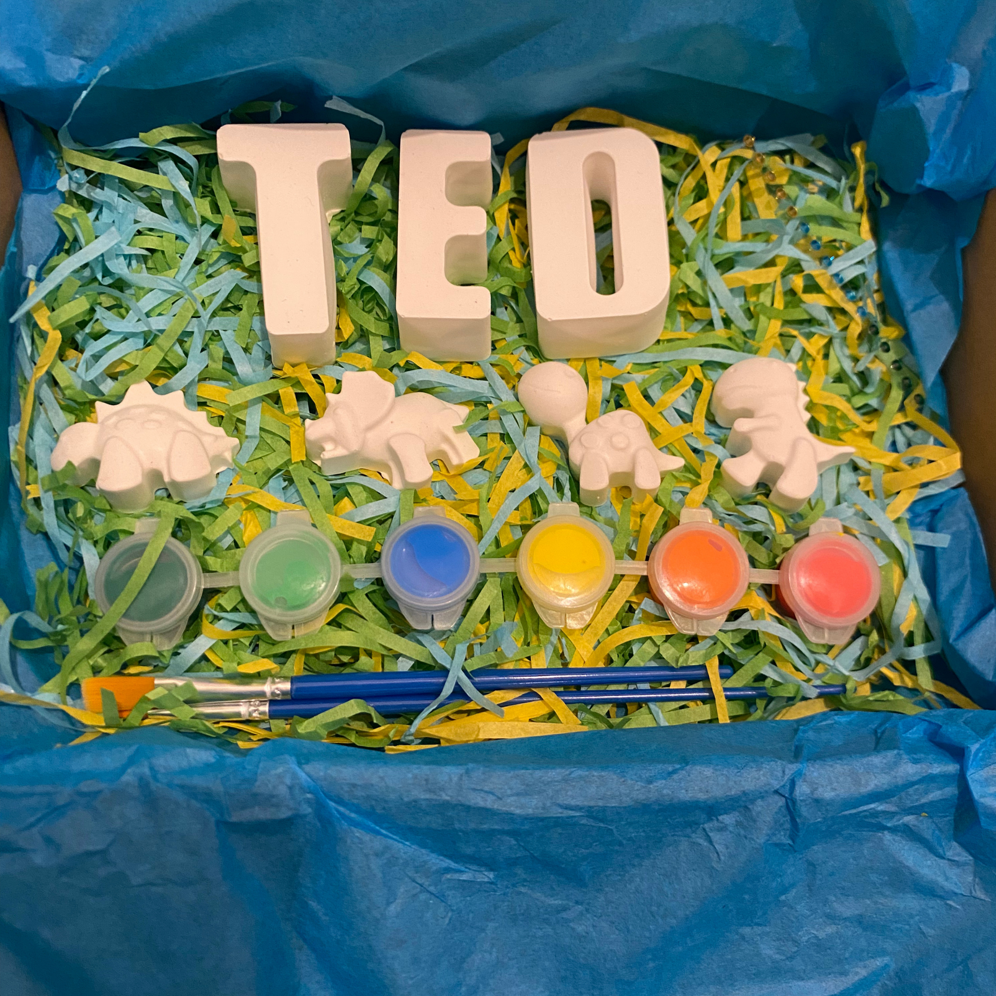 Children's craft kit with 'TED' letters, paint containers, and paintbrushes on a blue background.