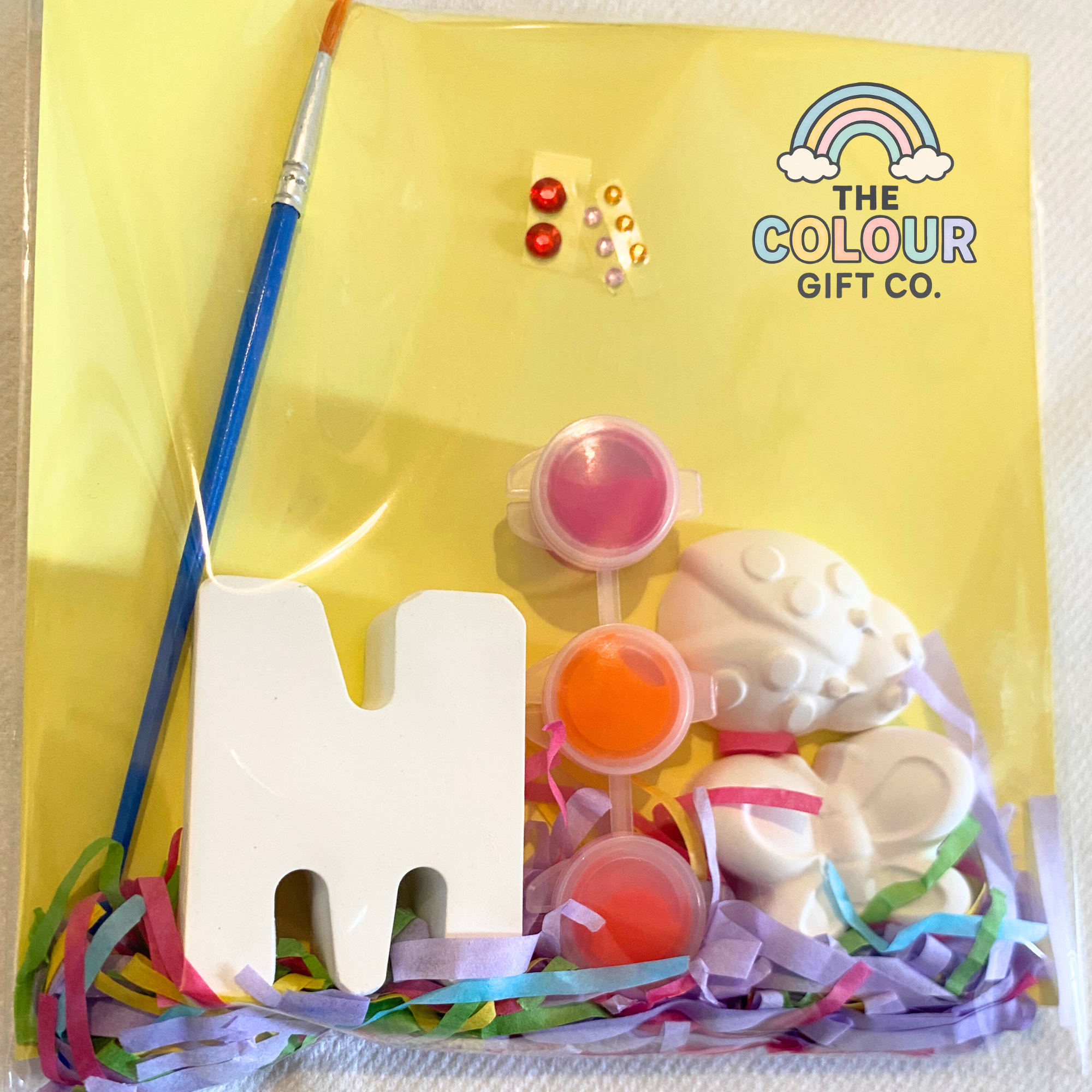 Colorful mini paint kit with letter 'M' and themed plaster mould on a yellow background with stickers, paint and brush with 'The Colour Gift Co.' branding.